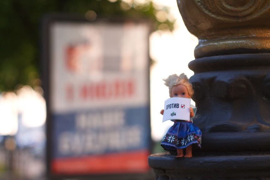 A miniature doll with blond hair and wearing a blue dress holds a piece of paper.