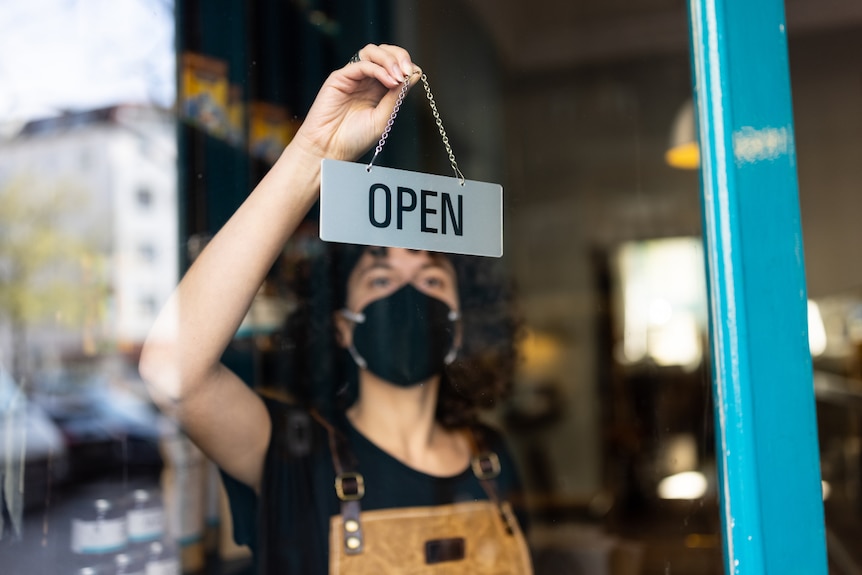 A young non-binary person hanging an open sign from the door of a small bakery.