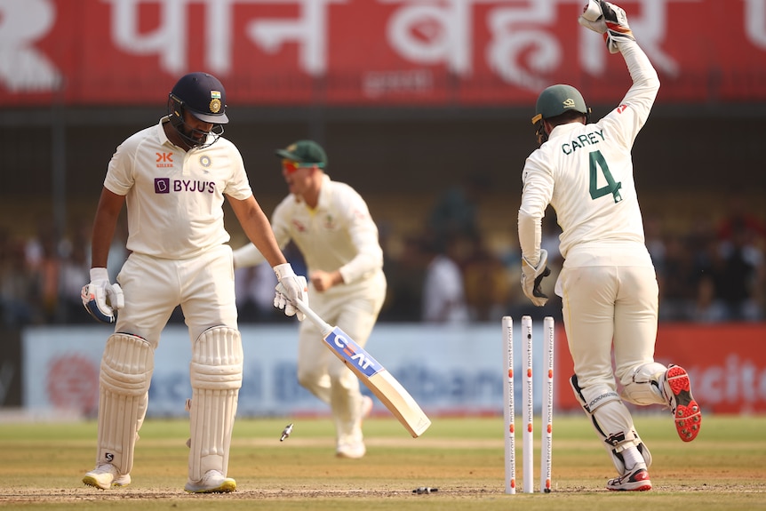 An Indian batsman dejectedly looks down at the stumps as the bails fly off and the wicketkeeper celebrates.