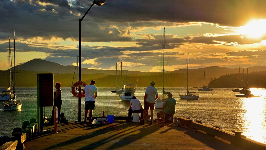 People partially silhouetted by the setting sun fish off the Eden wharf, with boats and yachts in the background.