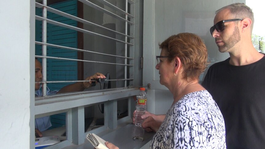A shorter woman talks to a man through a barred window while a taller man wearing sunglasses stands behind her.