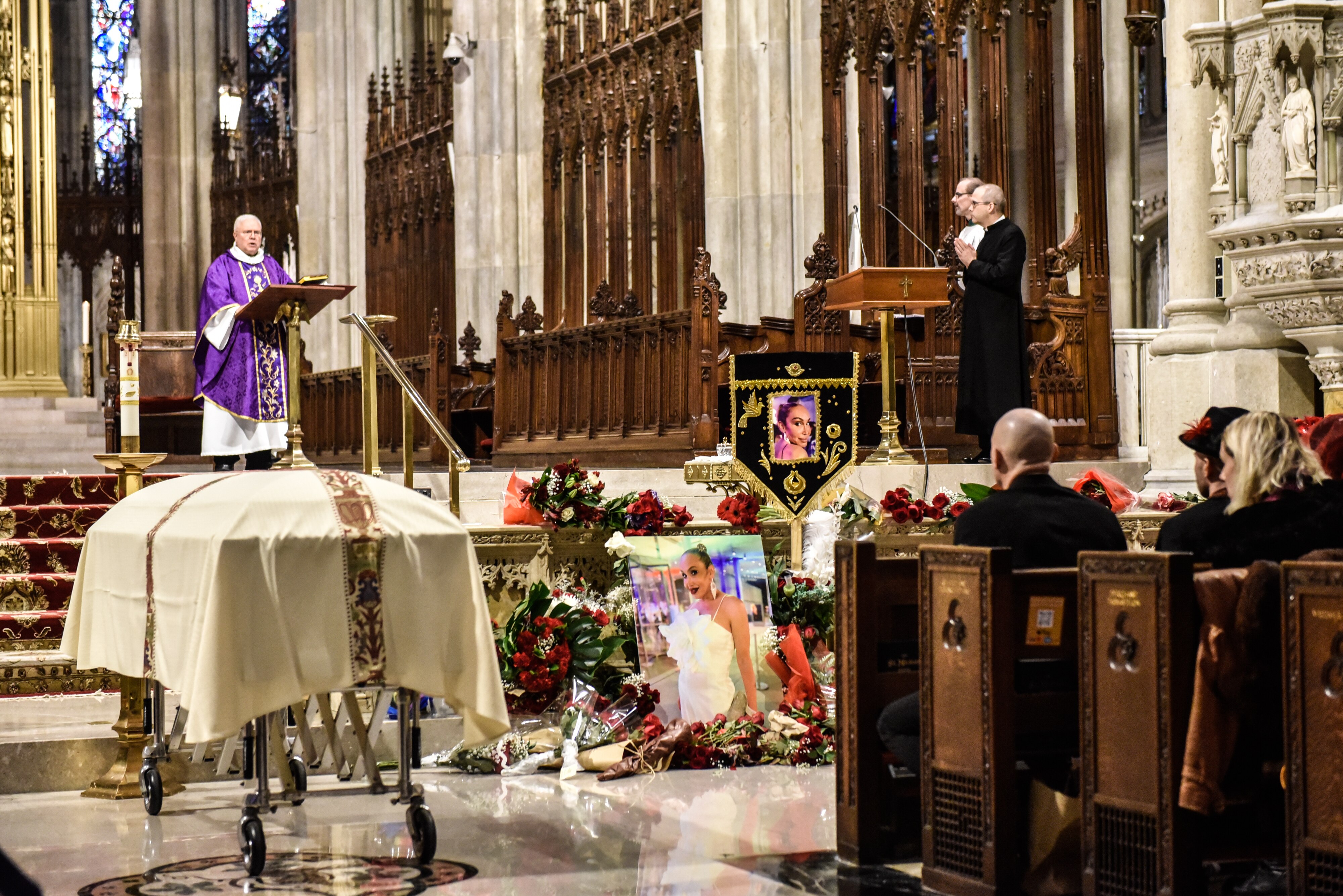 A wide shot of a priest speaking in a large church. People sitting in pews and standing nearby. A casket in the aisle.
