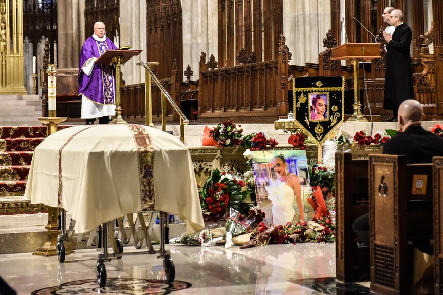 A wide shot of a priest speaking in a large church. People sitting in pews and standing nearby. A casket in the aisle.