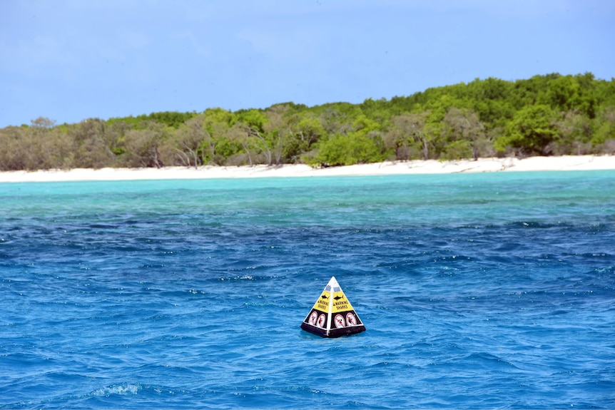 Different shades of blue and turquoise meet white sand, trees of island, triangle warning buoy floating in foreground.