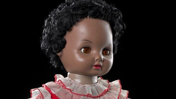 Manu the doll has brown skin and is reaching out. She wears a blue bottomed dress with a red top, has black hair and brown eyes.