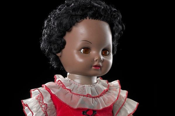 Manu the doll has brown skin and is reaching out. She wears a blue bottomed dress with a red top, has black hair and brown eyes.