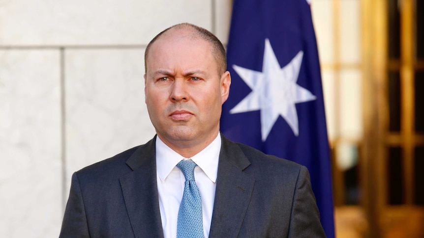 A man in a suit and tie speaking at a lecturn in front of a white marble wall with Australian flags in the background.