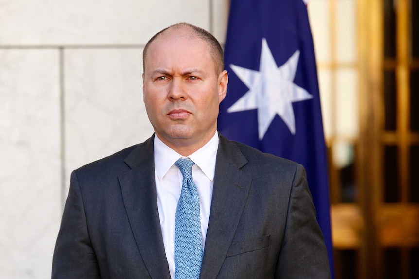 A man in a suit and tie speaking at a lecturn in front of a white marble wall with Australian flags in the background.