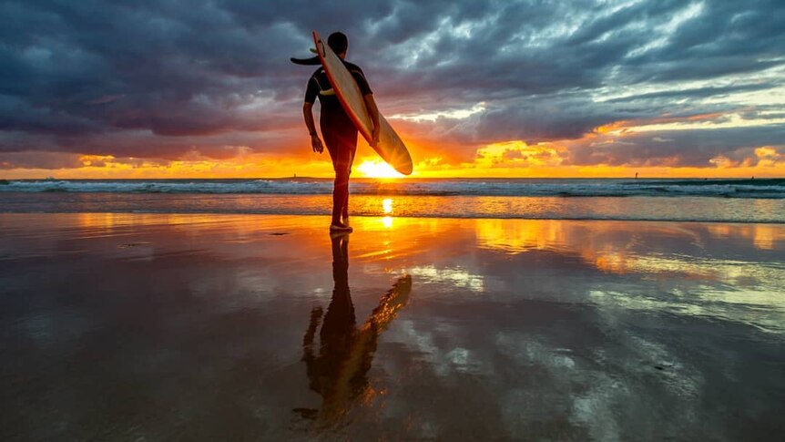 A person walks towards the water, carrying their surfboard as the sun rises.
