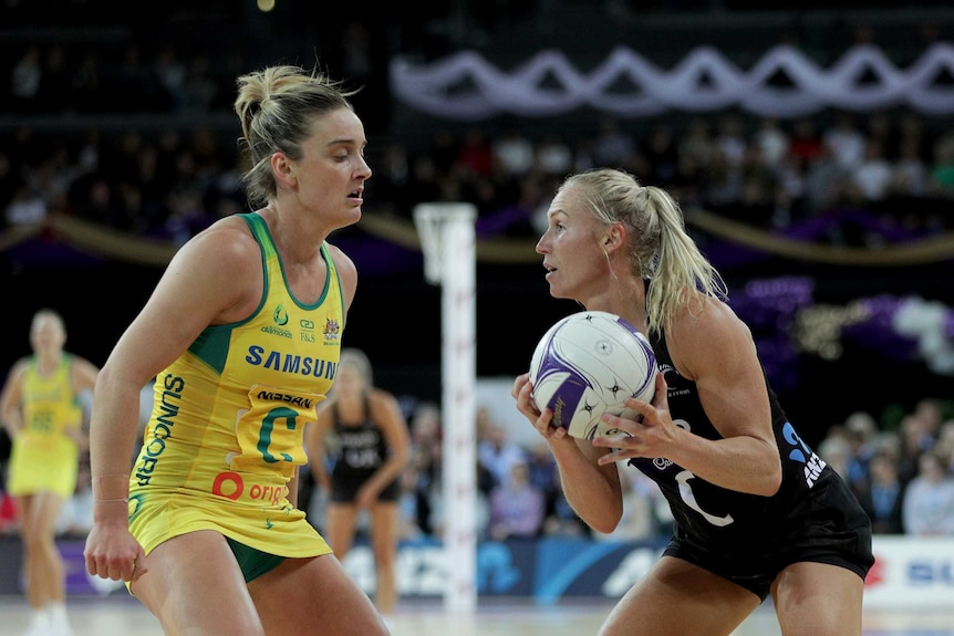 An Australian netballer stands in front of an New Zealand opponent holding the ball in the Constellation Cup Test.