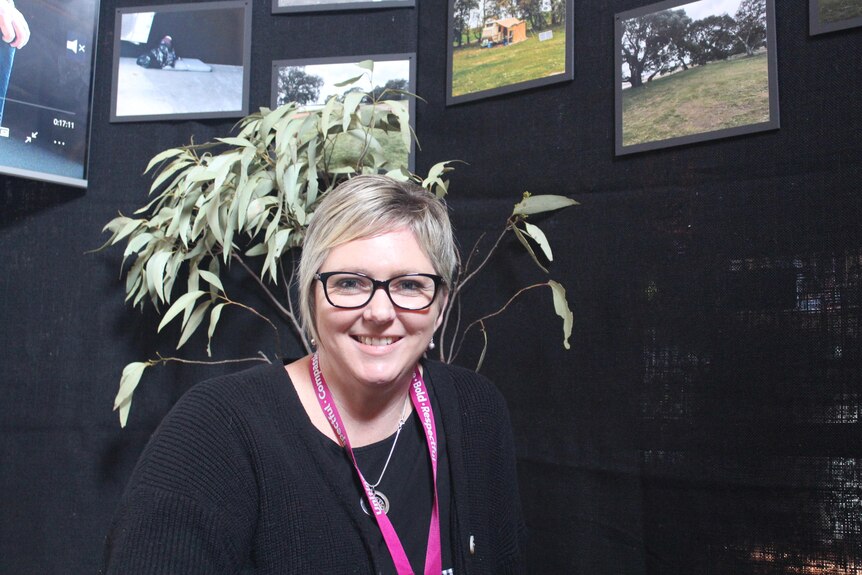 A woman sits in front of photos on a black wall and gum leaves. 