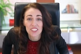 Jacinca Ardern leans against her desk towards the camera with an excited expression on her face