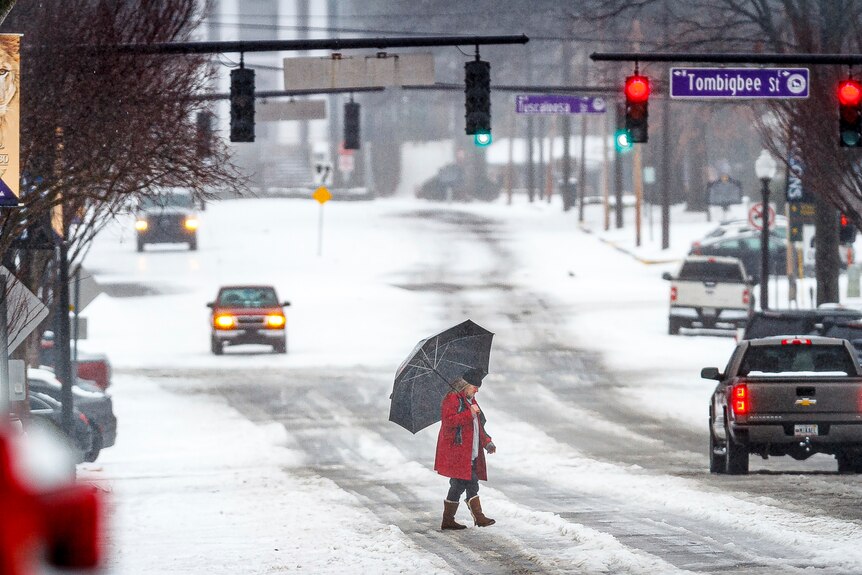 A man carrying an umbrella crosses a snowy road where a few cars are driving