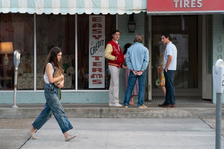 A long-haired brunette clutching a paper bag walks on the side of the street past a group of young men who stare at her