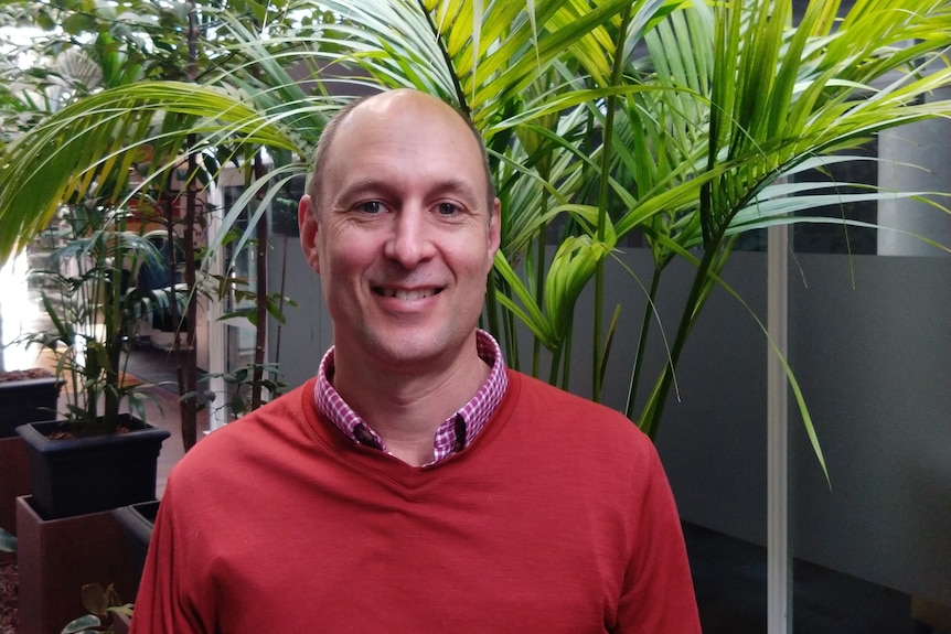 A man in a collared shirt and red jumper smiles at the camera.