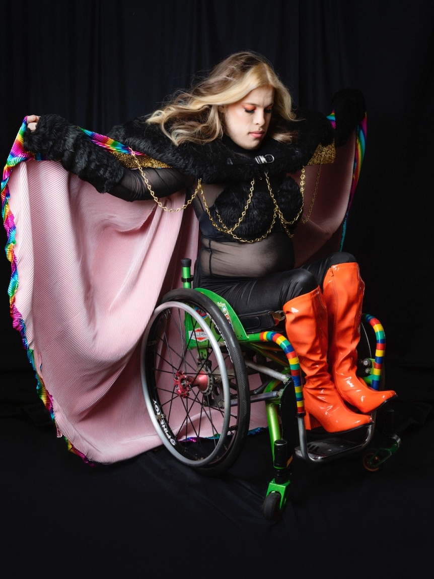 A person holding a rainbow coloured cape dressed in sheer black top, leather pants and orange knee-high boots in a wheelchair. 