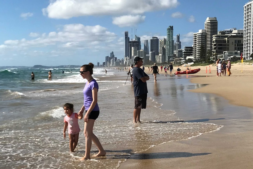 A woman in purple top holds hand of toddler in swimsuit on busy Gold Coast beach on a sunny morning.