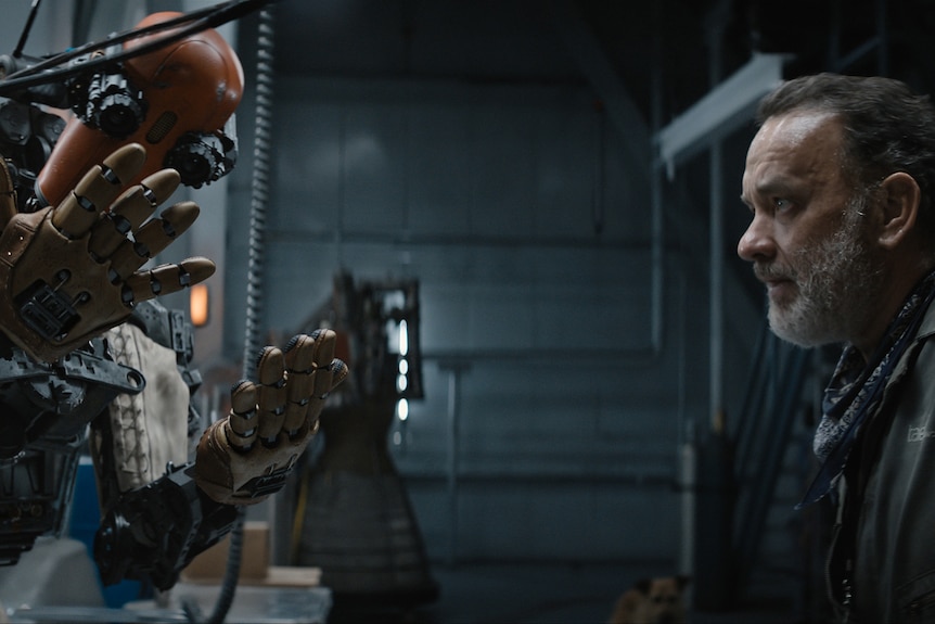 A metal human-shaped robot looks at its hands while a middle-aged man with grey beard looks on with interest bordering disbelief