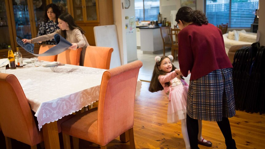 A girl in fluffy ears dances standing on her big sister's feet while another sister and the girls' mother set the dining table.