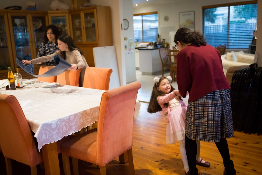 A girl in fluffy ears dances standing on her big sister's feet while another sister and the girls' mother set the dining table.