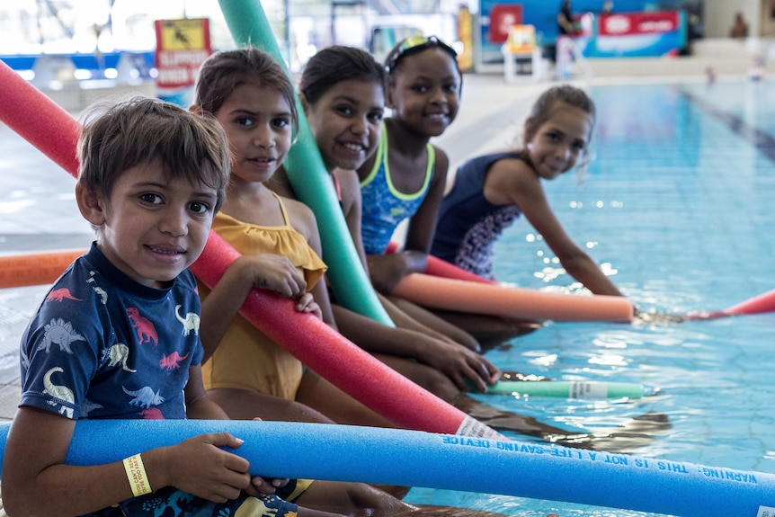 Kids with pool noodles sitting on the edge of an indoor swimming pool.
