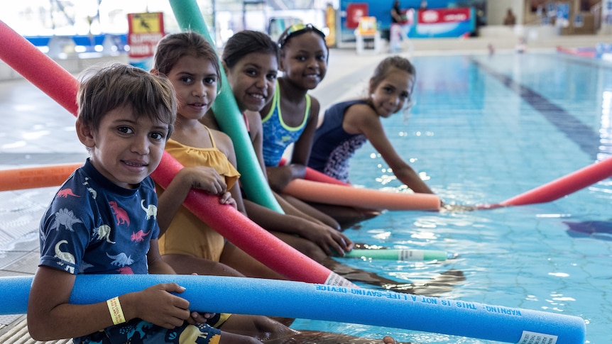 Kids with pool noodles sitting on the edge of an indoor swimming pool.