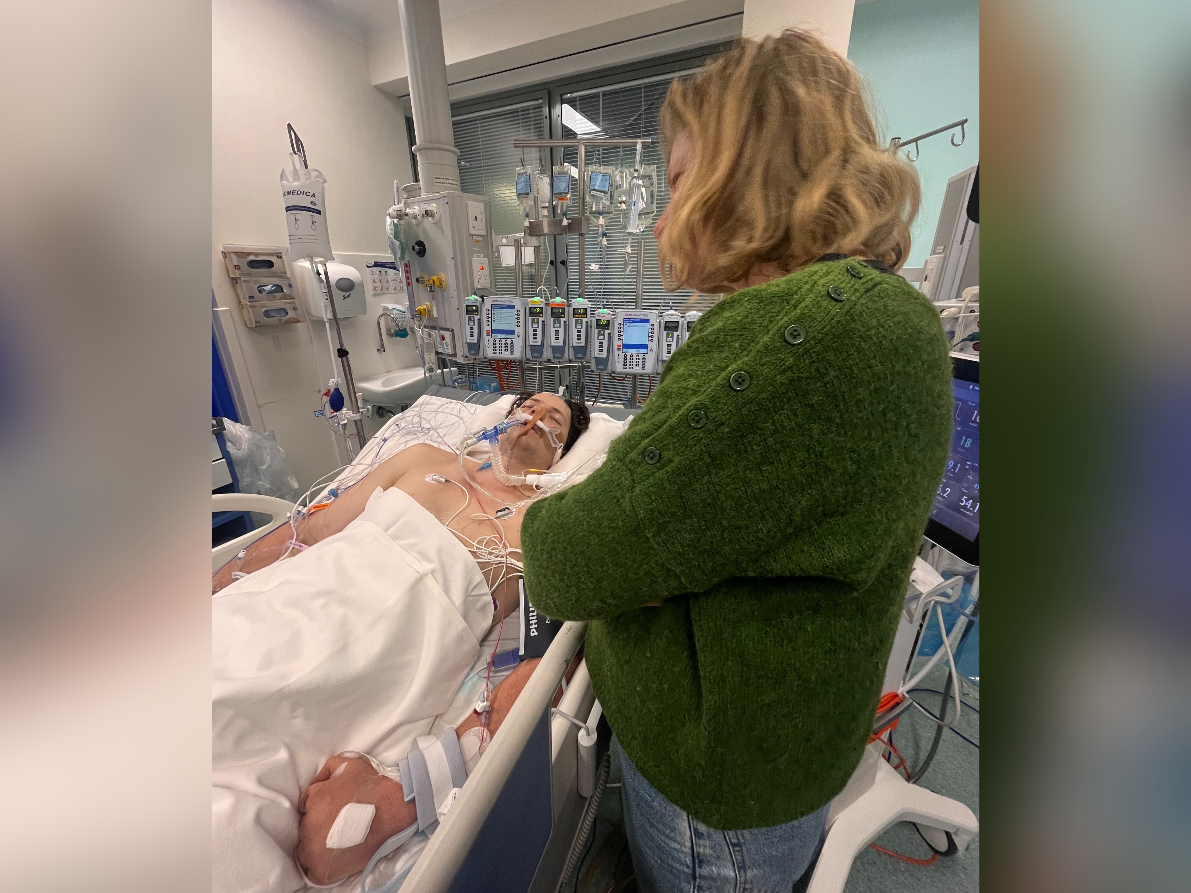 Xavier Sedger lying in a hospital bed with tubes in his nose. He wears a hospital gown.