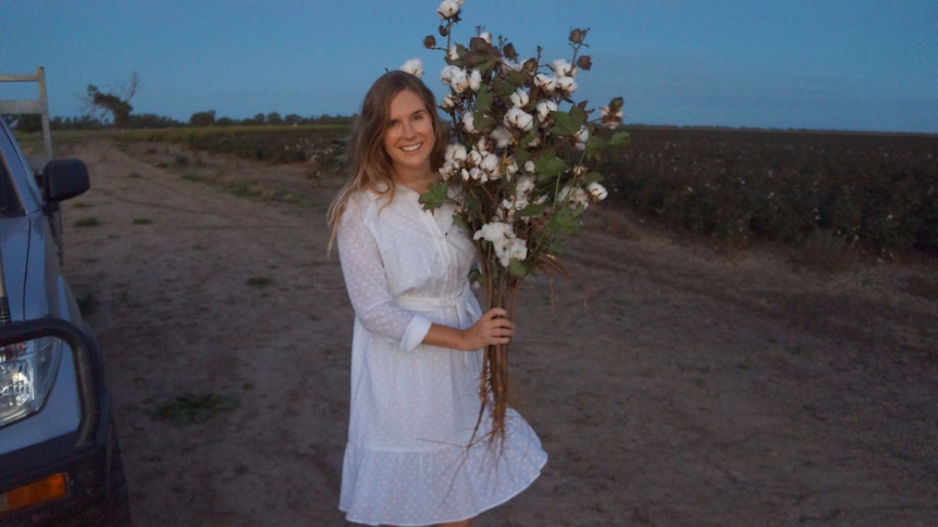 Emma Bond stands next to a cotton field holding a cotton flower bouquet, a ute is partially visible next to her.