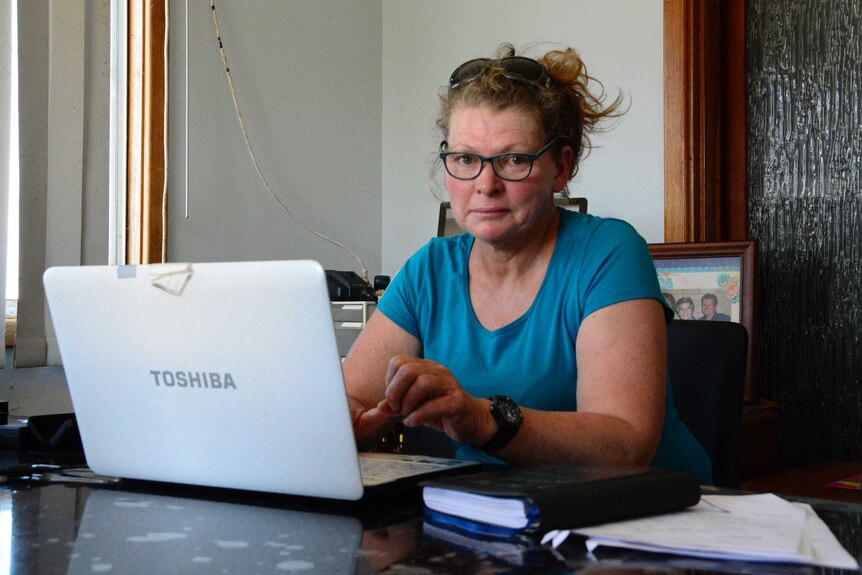 A woman sits indoors wearing a blue shirt and glasses posing for a photo at her laptop.