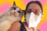 A selfie of a woman wearing a white mask and a fluffy ragdoll cat with blue eyes.