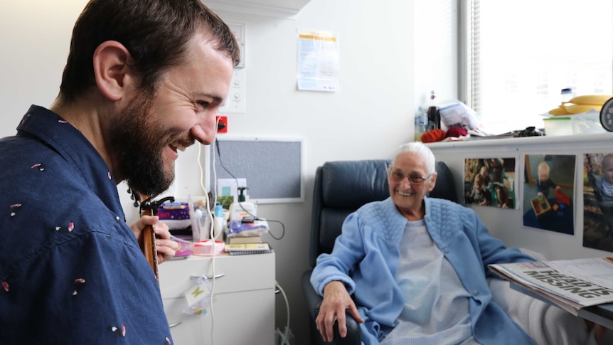 Will Parsell plays guitar to Marion Hayes in a hospital room in Hobart.