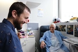 Will Parsell plays guitar to Marion Hayes in a hospital room in Hobart.