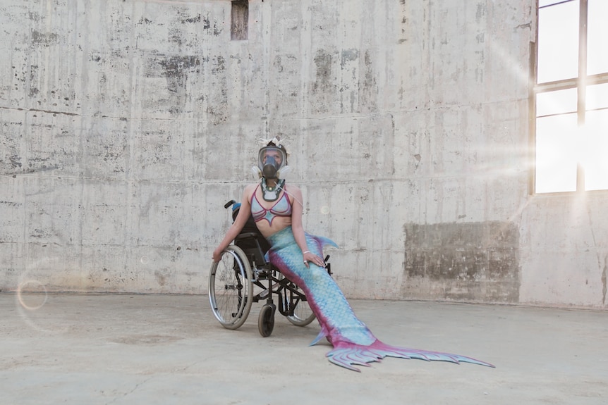 A woman in her mid 30s in a mermaid costume and oxygen mask sitting on a wheelchair, in a warehouse