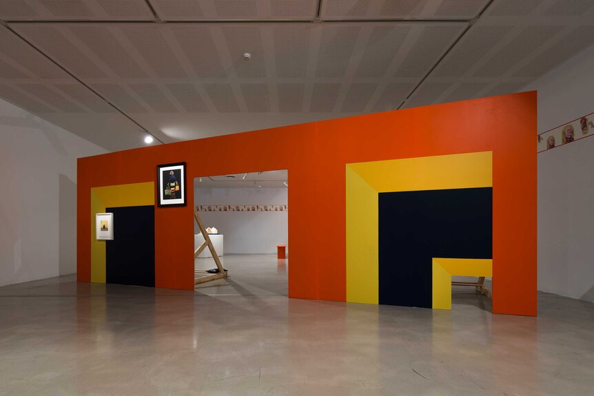 Colour photograph of a gallery space with bright coloured large scale wooden panels and assorted artworks.