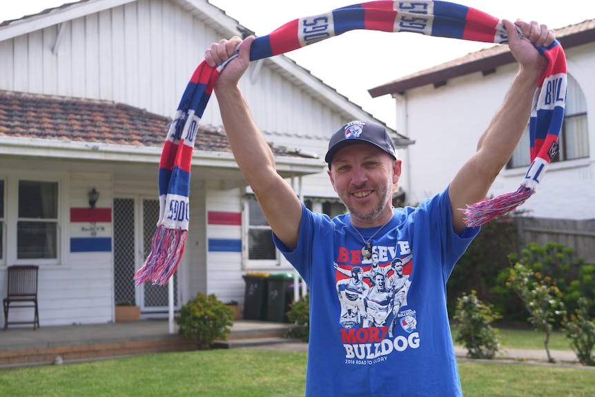 Shannon Yates stands in his front yard in a Bulldogs top, holding a team scarf in the air, in front on his painted house