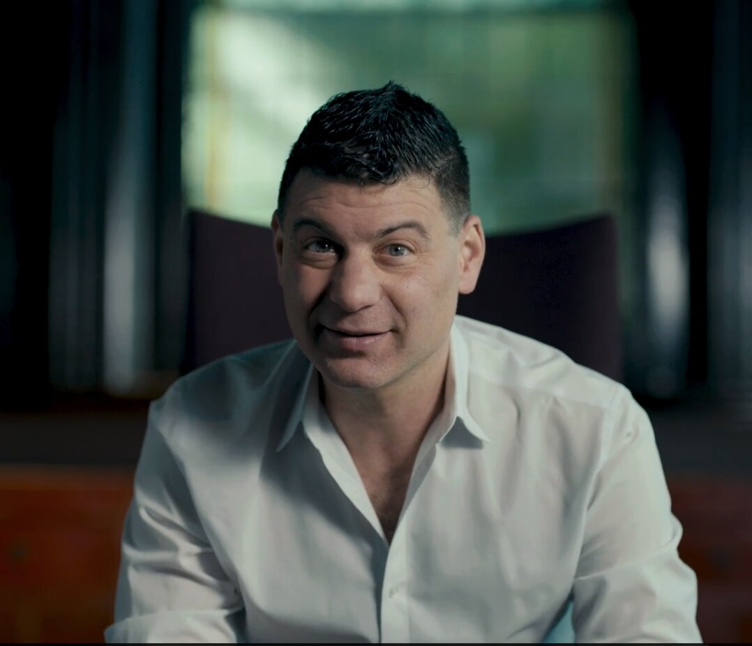 A dark haired man in a white collared shirt stares at the camera.
