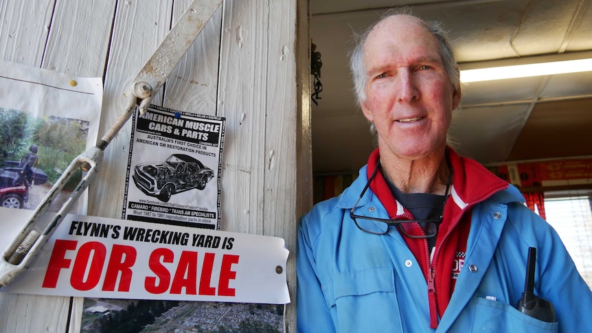 owner Wayne Flynn leans against office door with 'for sale' sign in the foreground