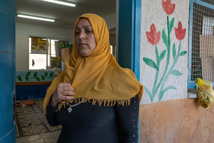 A Muslim woman in a yellow headscarf looks pensive standing in the doorway of a classroom