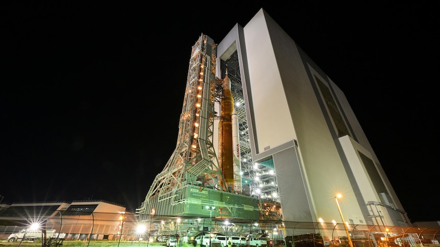 A rocket stands alongside a vertical building and scaffolding at night time 