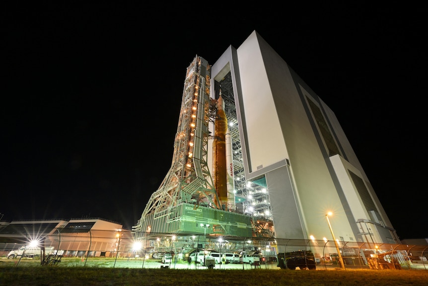 A rocket stands alongside a vertical building and scaffolding at night time 