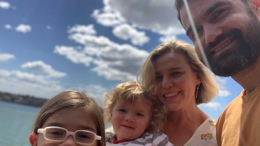 A man and woman and their two children smile to camera, with the beach and clouds in the distance.