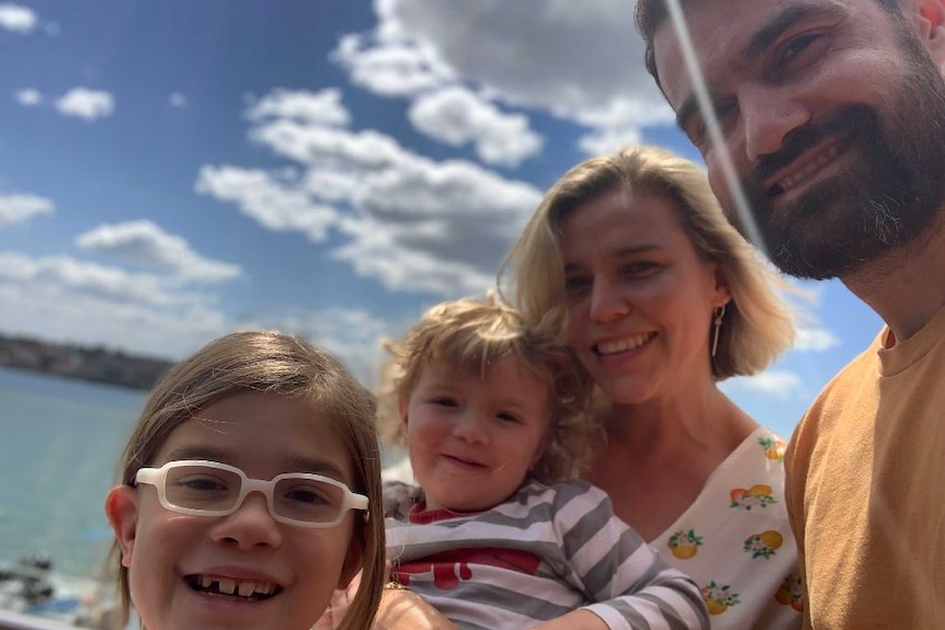 A man and woman and their two children smile to camera, with the beach and clouds in the distance.