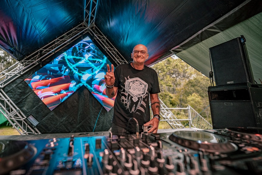 Photo of older man making peace sign standing behind DJ decks
