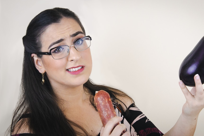 Nicola Macri holding an eggplant and salami with a puzzled look on her face. She wears glasses and has long black hair.
