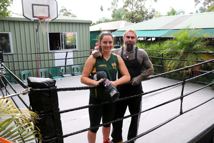 A woman holding boxing gloves stands in a backyard boxing ring with a heavily tattooed man with his arm around her.