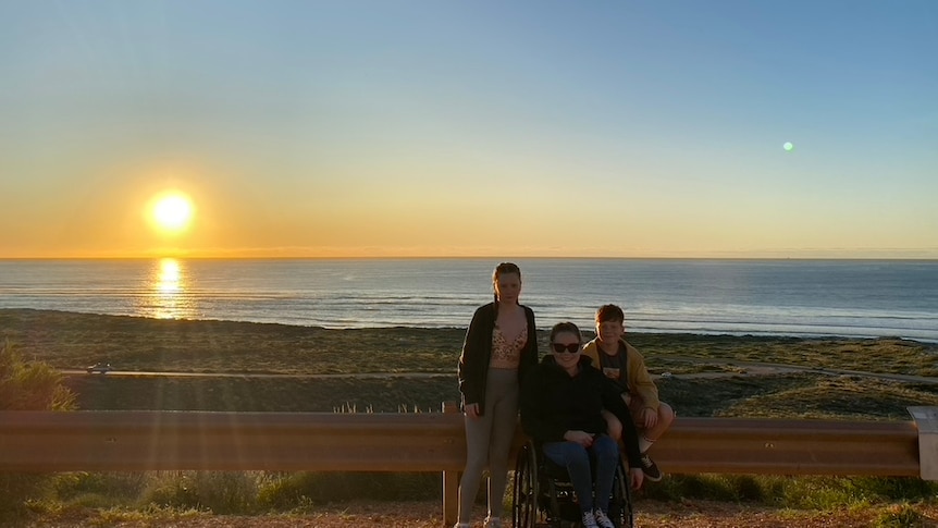 A family of three walk along a beach as the sun sets behind them