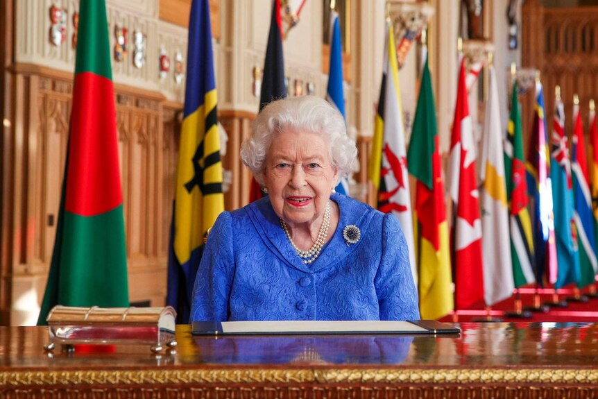 Queen Elizabeth sits at a desk in a large, opulent room with a large number of flags behind her.