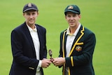 Two captains in blazers and caps stand together on the ground holding the Ashes.