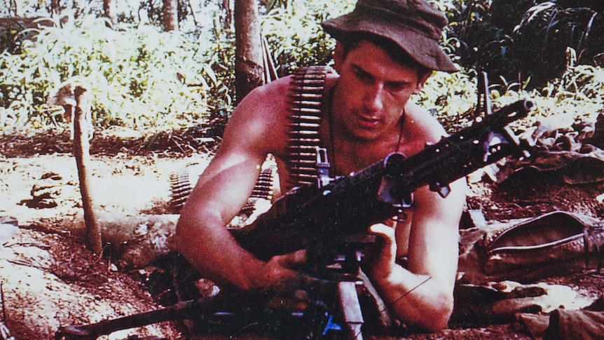 A shirtless John Wells lies in the dirt as he loads a machine gun.