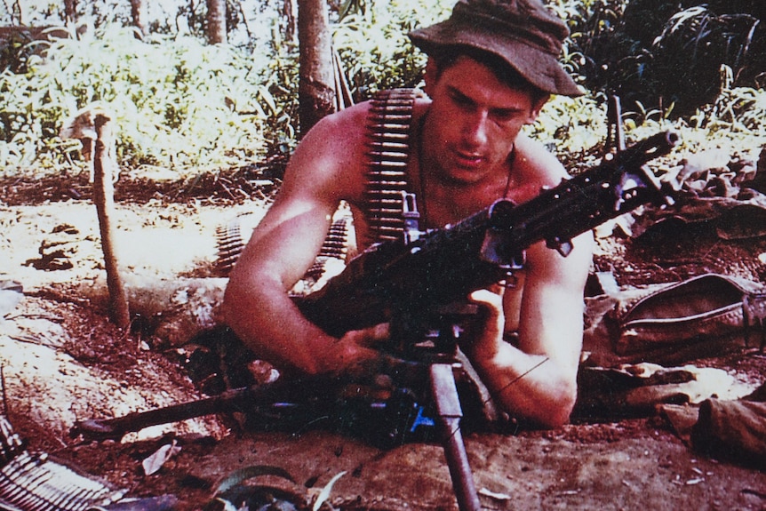 A shirtless John Wells lies in the dirt as he loads a machine gun.